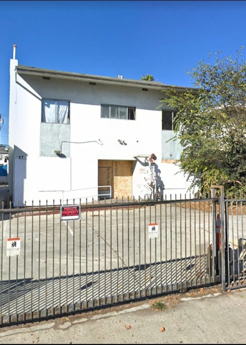 Building before — white modernist home with boarded windows and security fence